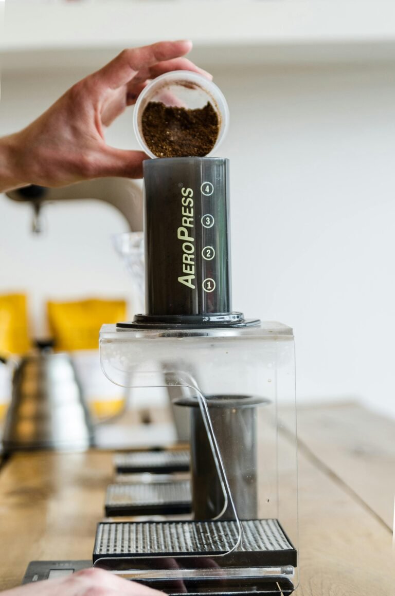 A hand pouring coffee grounds into an Aeropress, set on a table with a blurred background.